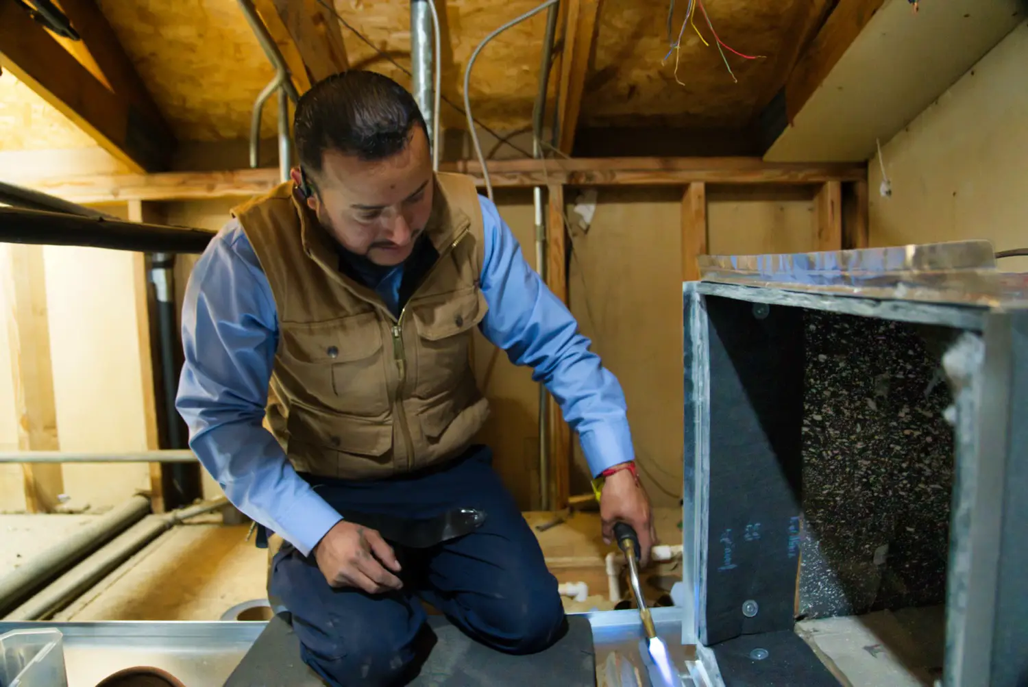 Nalk technician kneeling in an attic beside an indoor HVAC unit with its filter compartment open
