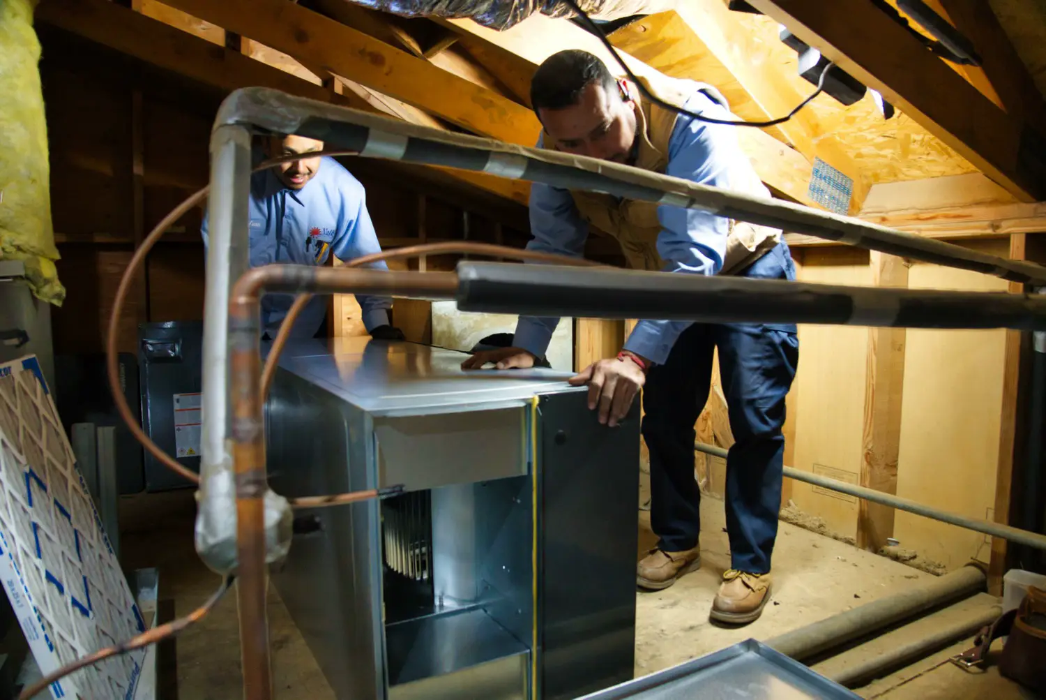Two Nalk technicians working around an indoor air handler in an attic